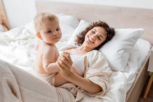 Mujer feliz en ropa de salón acostada en la cama y cogida de la mano de la hija lactante - foto de stock