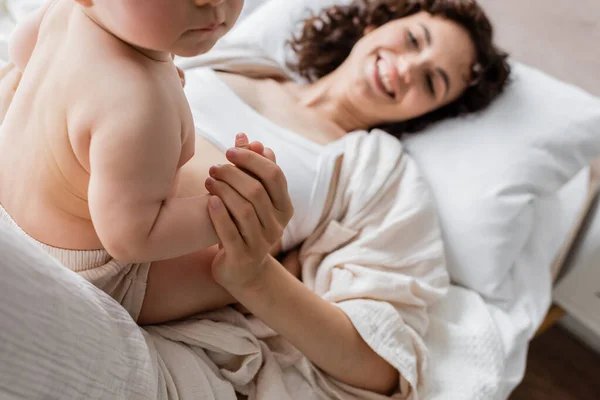 Весела жінка в лаунж-одязі лежить на ліжку і тримає руку немовляти дочки — стокове фото