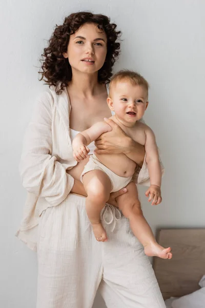 Brunette woman in pajamas holding baby and looking at camera at home — Stock Photo