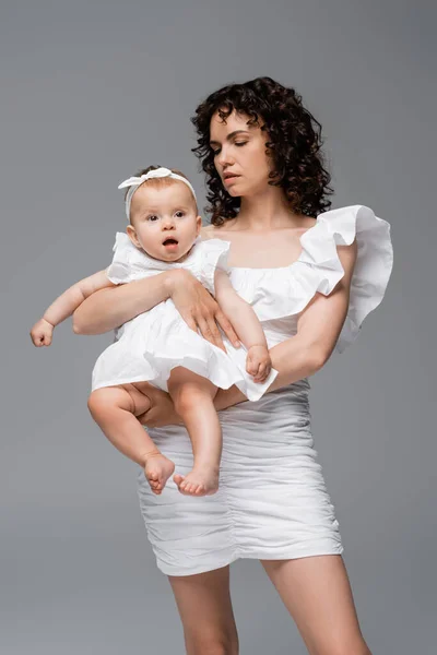 Stylish mother in white dress holding baby daughter isolated on grey — Stock Photo