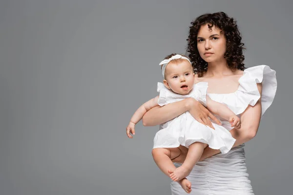 Mujer rizada en vestido blanco sosteniendo niña aislada en gris - foto de stock