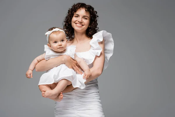 Mãe sorridente em vestido branco elegante segurando bebê menina isolada no cinza — Fotografia de Stock