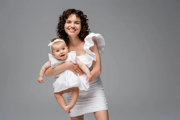 Cheerful brunette woman in dress holding happy baby daughter isolated on grey — Stock Photo