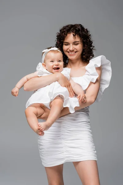 Brünette Frau im Kleid hält fröhliche kleine Tochter isoliert auf grau — Stockfoto