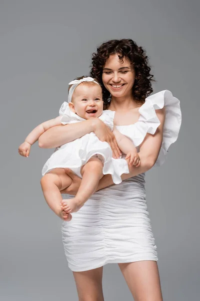 Mulher morena no vestido segurando alegre bebê filha isolada no cinza — Fotografia de Stock