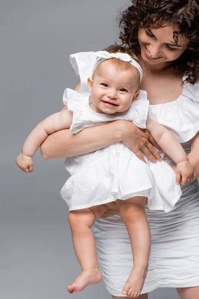 Sonriente mamá sosteniendo elegante bebé hija en vestido y diadema aislado en gris - foto de stock