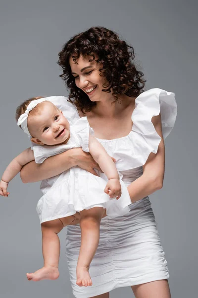 Sonriente madre mirando linda niña en vestido aislado en gris - foto de stock