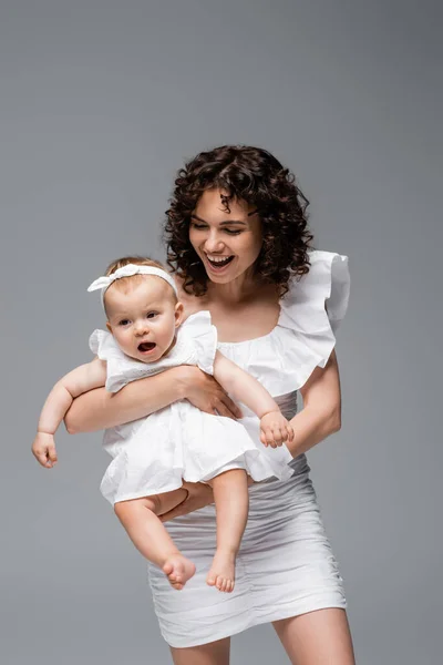 Позитивная кудрявая мама держит маленькую дочь в платье, изолированном от серых — стоковое фото
