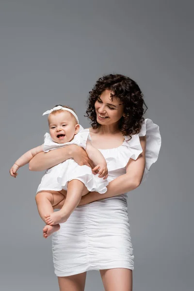 Smiling curly mother holding cheerful baby in dress isolated on grey — Stock Photo