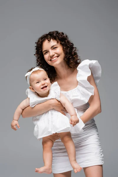 Brunette woman looking at camera while holding baby in dress isolated on grey — Stock Photo