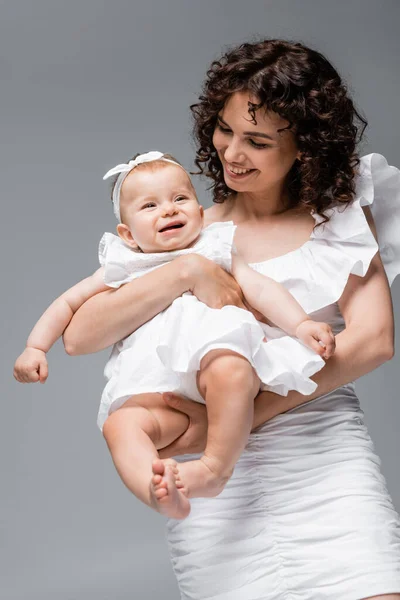 Stylish mom looking at smiling baby girl in white dress and headband isolated on grey — Stock Photo