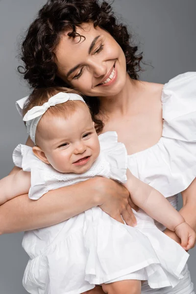 Joyful woman hugging smiling baby daughter in dress isolated on grey — Stock Photo