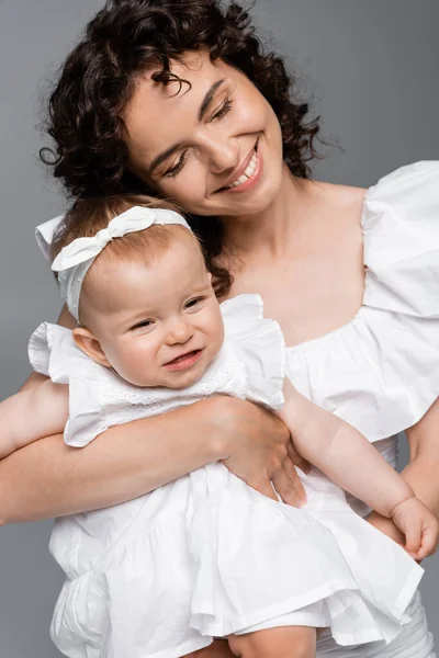 Mulher alegre abraçando bebê sorridente filha no vestido isolado no cinza — Fotografia de Stock