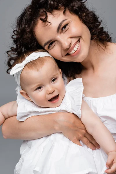 Madre positiva mirando a la cámara mientras sostiene al niño en vestido aislado en gris - foto de stock