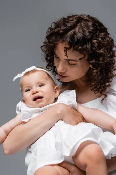 Curly parent looking at baby girl in white headband and dress isolated on grey — Stock Photo