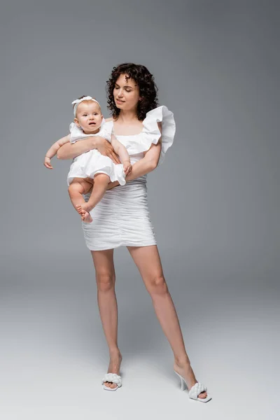 Moda mamá en vestido y tacones celebración bebé hija en gris fondo - foto de stock