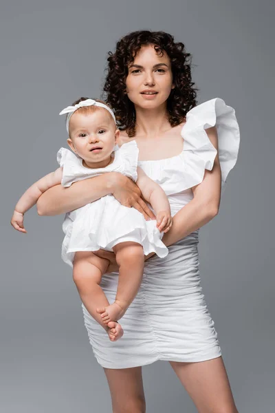 Trendige Mutter im Kleid blickt in die Kamera, während sie das kleine Mädchen isoliert auf grau hält — Stockfoto