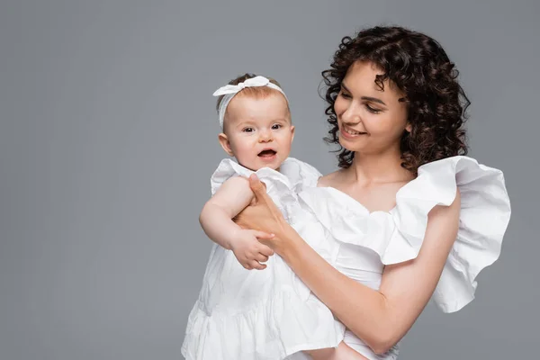 Curly woman holding cheerful baby girl in dress isolated on grey — Stock Photo