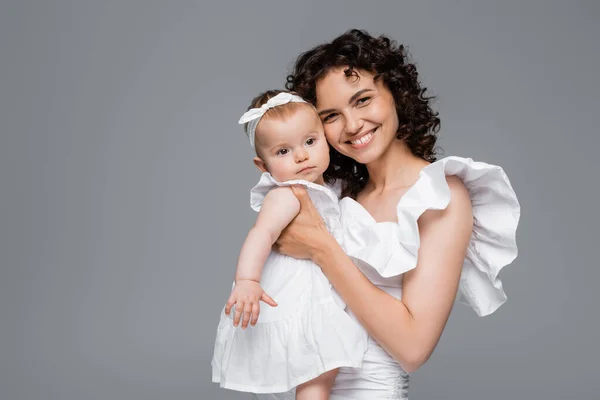Lächelnde Mutter blickt in die Kamera, während sie das kleine Mädchen in weißer Kleidung isoliert auf grau hält — Stockfoto