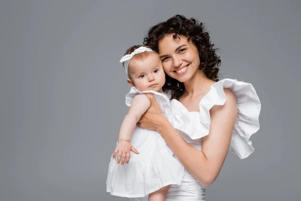 Sonriente mamá mirando a la cámara mientras sostiene a la niña en ropa blanca aislada en gris - foto de stock