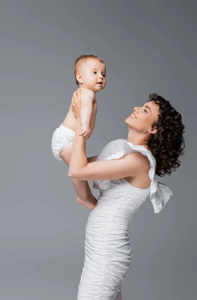 Mãe encaracolado feliz no vestido segurando bebê filha isolada no cinza — Fotografia de Stock