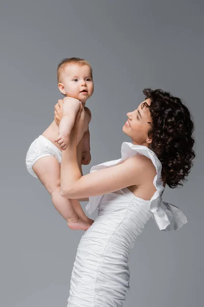 Elegante mamma in abito bianco che tiene il bambino in mutandine isolato sul grigio — Foto stock