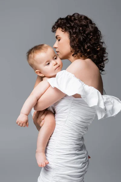 Mujer rizada en vestido abrazando hija bebé aislado en gris - foto de stock