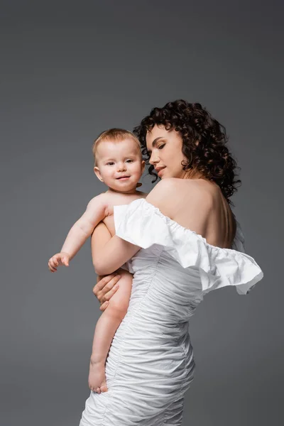 Brunette woman in dress holding smiling infant daughter isolated on grey — Stock Photo