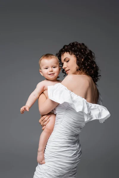 Mulher morena em vestido segurando sorridente criança filha isolada em cinza — Fotografia de Stock