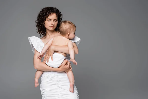 Elegante mamá en vestido blanco abrazando a la hija en bragas aisladas en gris - foto de stock