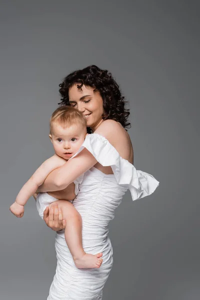 Happy woman in stylish white dress hugging infant child isolated on grey — Stock Photo