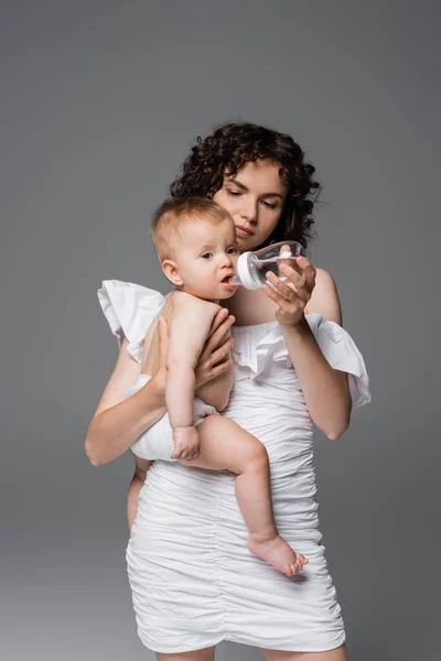 Stylish mom holding baby bottle and infant daughter isolated on grey — Stock Photo