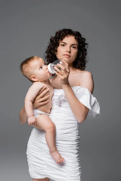 Mulher na moda em vestido branco segurando filha e mamadeira enquanto olha para a câmera isolada no cinza — Fotografia de Stock