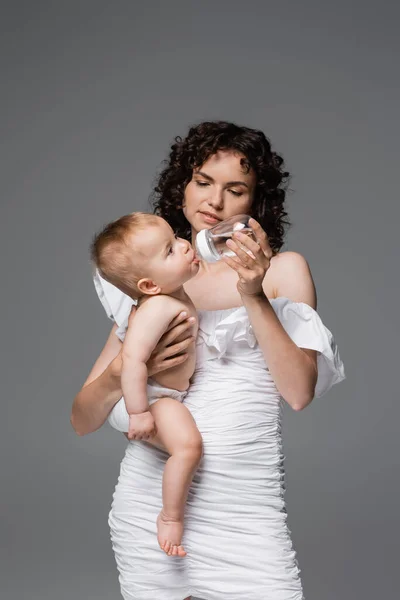 Maman souriante en robe tendance tenant biberon avec de l'eau près de la fille en culotte isolée sur gris — Photo de stock