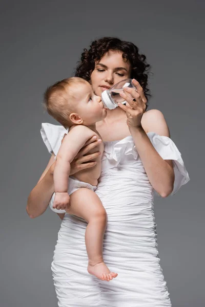 Trendy woman in dress holding baby and bottle with water isolated on grey — Stock Photo