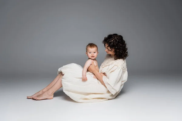 Lockige Mutter im Kleid hält lächelndes braunes Mädchen, während sie auf grauem Hintergrund sitzt — Stockfoto