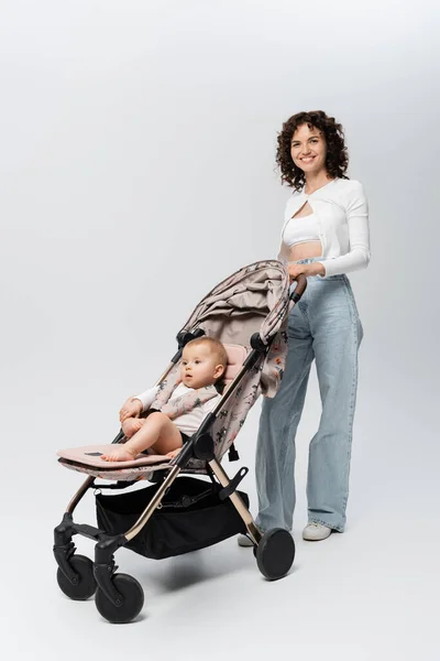 Piena lunghezza di madre sorridente guardando la fotocamera vicino al bambino in passeggino su sfondo grigio — Foto stock