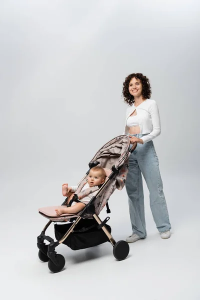 Curly pai de pé perto da filha infantil no carrinho de bebê em fundo cinza — Fotografia de Stock