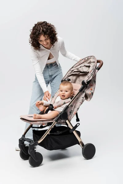 Smiling woman looking at baby daughter in stroller on grey background — Stock Photo