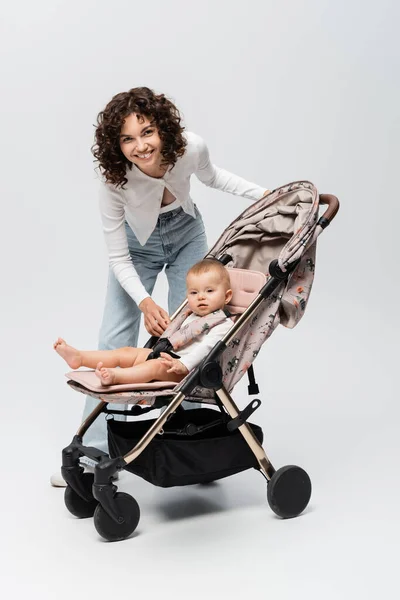 Felice madre guardando la macchina fotografica vicino alla bambina in passeggino su sfondo grigio — Foto stock