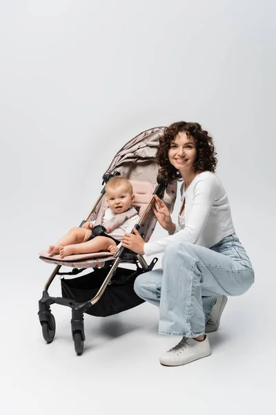 Smiling parent and baby girl in stroller looking at camera on grey background — Stock Photo