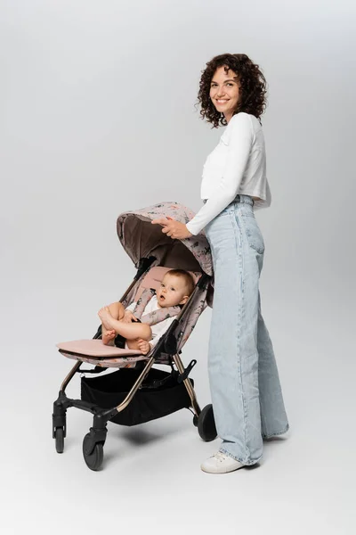 Madre riccia in jeans in piedi vicino alla bambina in passeggino su sfondo grigio — Foto stock