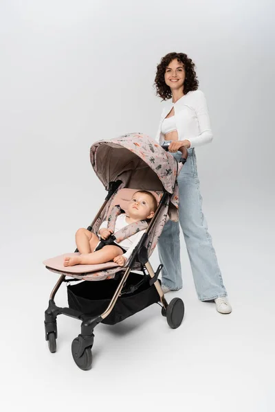 Baby-Mädchen sitzt im Kinderwagen neben lächelnder Mutter und blickt in die Kamera auf grauem Hintergrund — Stockfoto