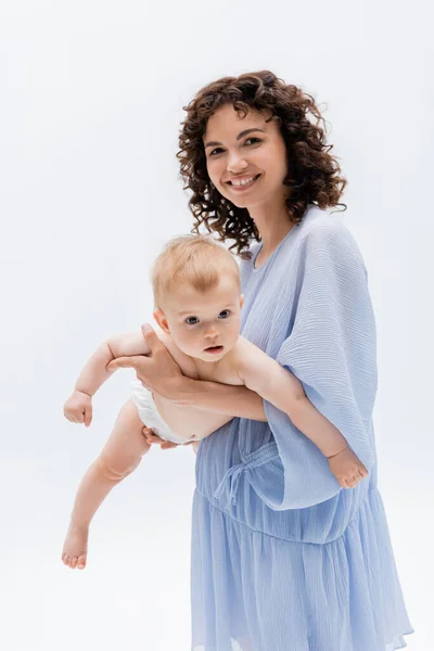 Mujer rizada en vestido azul sosteniendo a la hija lactante en bragas aisladas en blanco - foto de stock