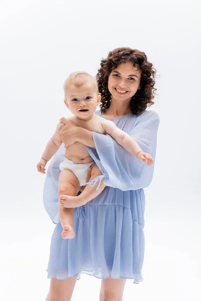 Donna in abito blu abbracciare il bambino in mutandine e guardando la fotocamera isolata su bianco — Foto stock