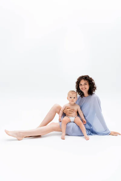 Smiling woman looking at camera while holding baby girl and sitting on white background — Stock Photo