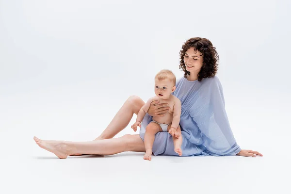 Curly woman in dress looking at infant daughter while sitting on white background — Stock Photo