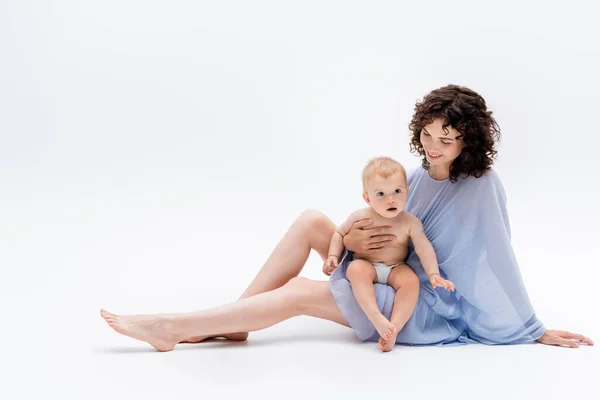 Maman agréable en robe bleue touchant bébé fille tout en étant assis sur fond blanc — Photo de stock