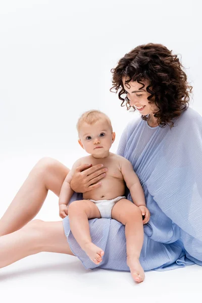 Positivo morena mamá mirando bebé hija mientras sentado en blanco fondo - foto de stock