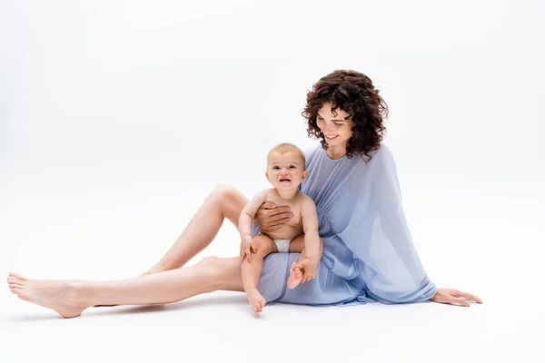 Barefoot woman in blue dress looking at happy baby girl on white background — Stock Photo